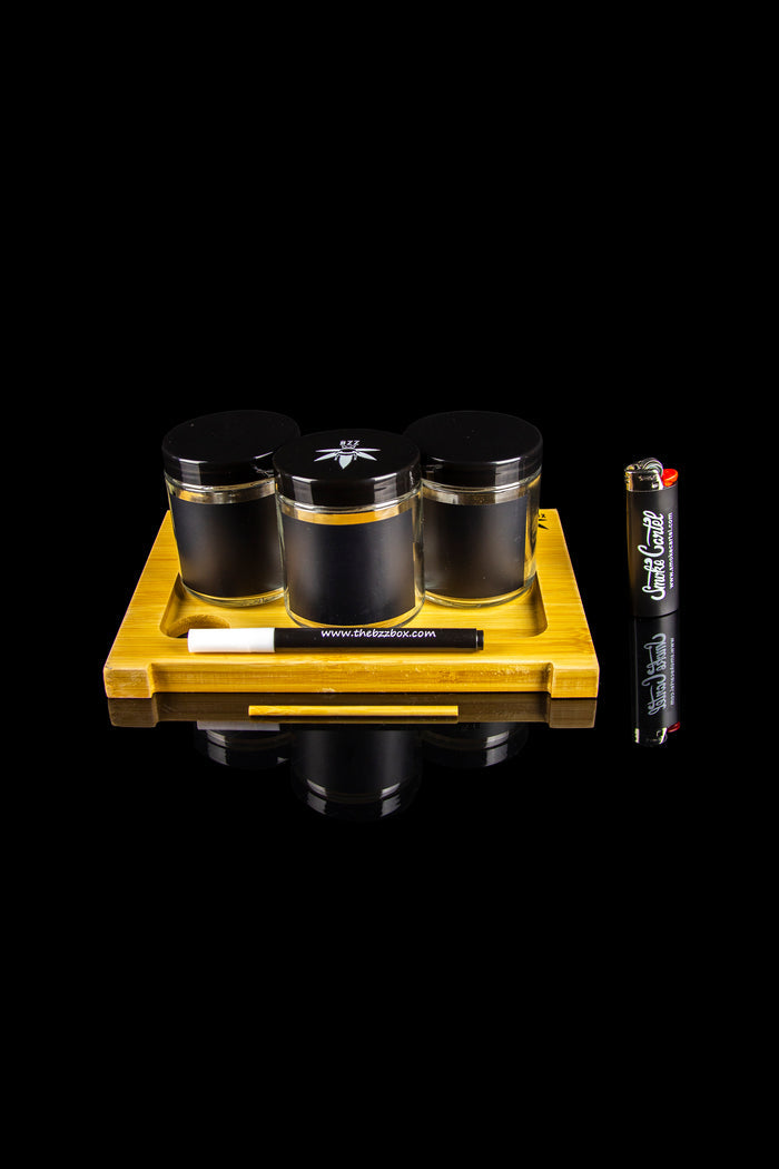 Cannabis storage jars, a grinder, rolling papers, and a lighter arranged on a wooden display tray.