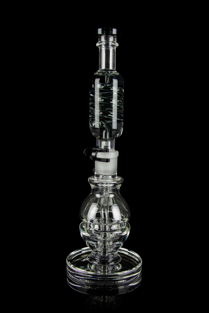 Clear glass water pipe (rig) with intricate bubble and cylinder design, reflecting on a black surface.