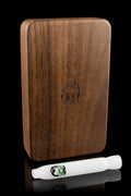 Wooden cannabis smoking case with engraved skull design, alongside a white one-hitter pipe on a reflective surface.